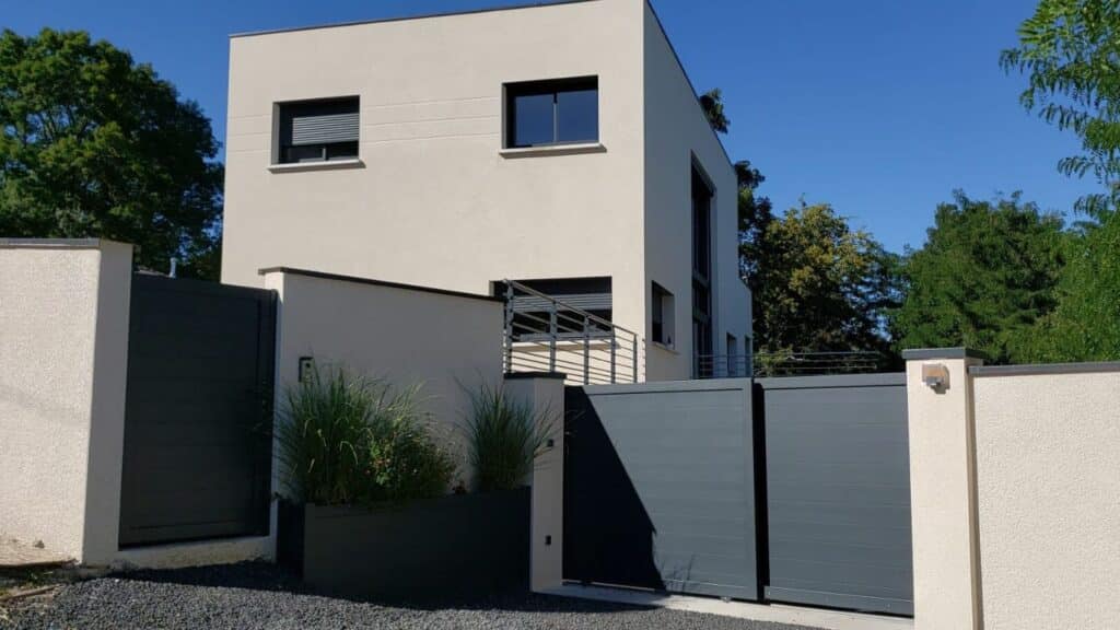 Maison moderne : façade beige, portail anthracite et design. Architecture moderne beige avec cadres de fenêtres sombres. Mur d'enceinte et portails coulissants anthracite devant un jardin minimaliste.